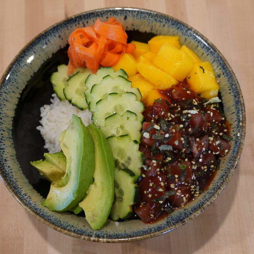 A colorful poke bowl with tuna, mango, avocado, cucumber, pickled ginger, and rice.