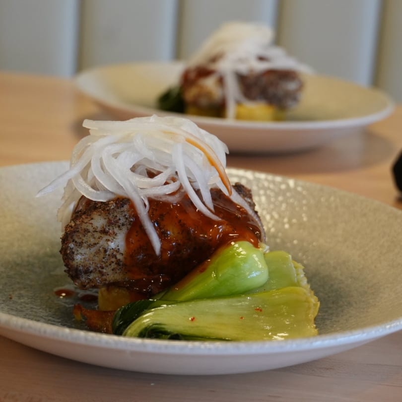 Two plates of meat with red sauce, sliced onions, and bok choy on a wooden table.