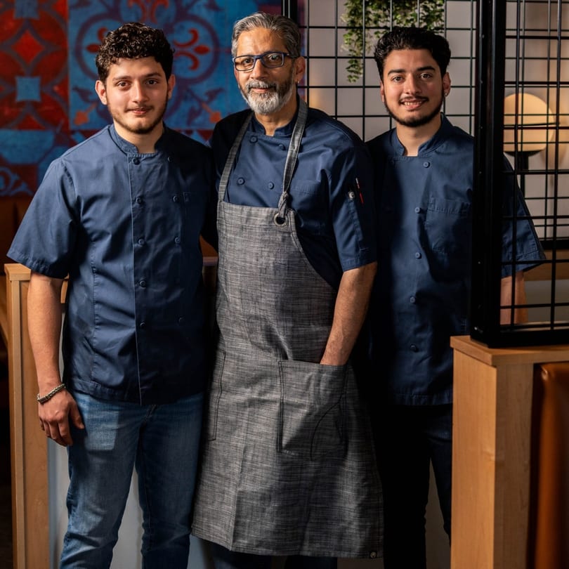 Three male chefs in blue uniforms, one with a grey apron, stand smiling in a restaurant.
