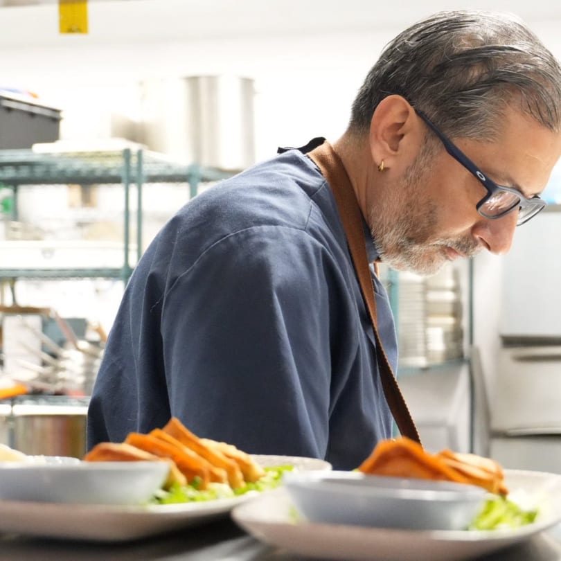 Chef in blue uniform and glasses plating food with fries and salad in a commercial kitchen.