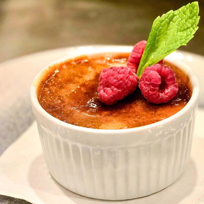 Crème brûlée in a white ramekin, garnished with fresh raspberries and a mint leaf.
