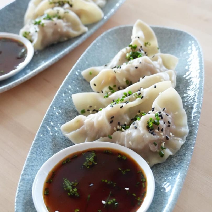 Two plates of PanAsian dumplings, garnished with sesame seeds and chives, with dipping sauce.