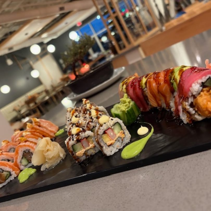 A black platter holds three types of sushi rolls, with wasabi and pickled ginger, in a restaurant.